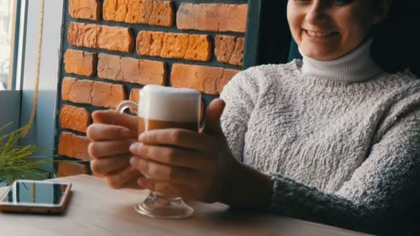 Güzel kız ikindilerde içecekler kahve latte hava köpük ile gülümseyen rahat kafede oturur ve pencereden görünüyor — Stok video