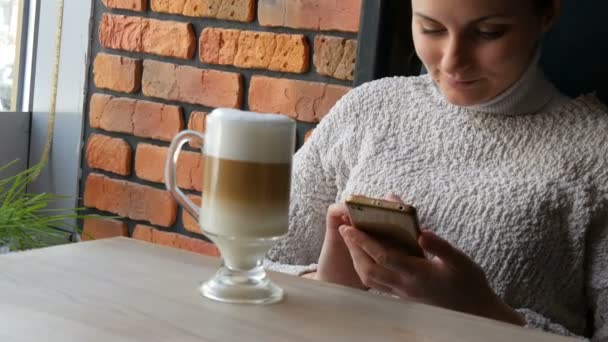 Красивая девушка сидит в уютном кафе задумчиво улыбаясь пьет кофе латте с пеной воздуха и смотрит в окно — стоковое видео