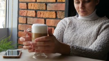 Güzel kız ikindilerde içecekler kahve latte hava köpük ile gülümseyen rahat kafede oturur ve pencereden görünüyor