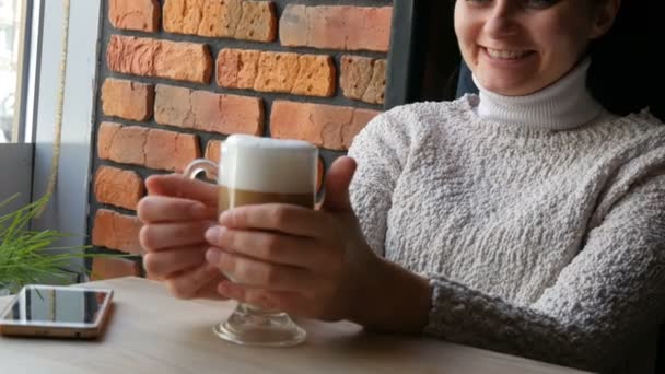 Mooi meisje zit in het gezellige café pensively glimlachend dranken koffie latte met lucht schuim en kijkt uit het raam — Stockvideo