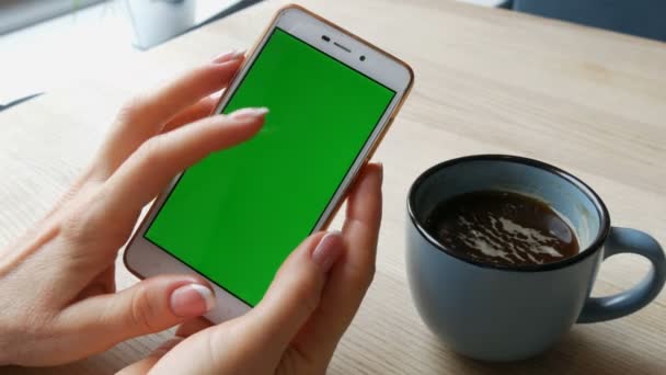 Smartphone de pantalla verde. Chroma Key en un teléfono inteligente blanco, las manos femeninas sostienen el teléfono móvil en un café al lado de una taza de café — Vídeos de Stock