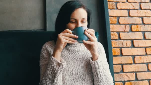 Hermosa chica se sienta en un acogedor café sonriendo pensativamente bebe café y mira por la ventana — Vídeo de stock