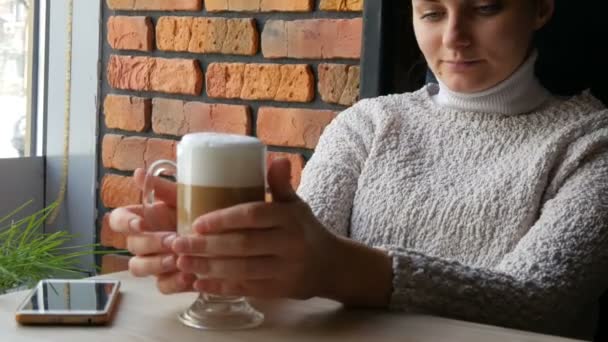 Vacker flicka sitter i mysiga kafé tonföljd leende drycker kaffe latte med air skum och ser ut genom fönstret — Stockvideo