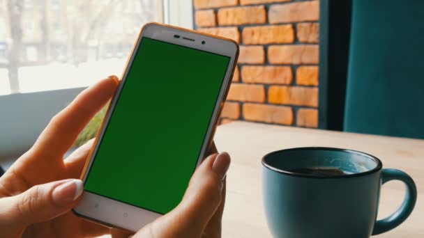 Smartphone de pantalla verde. Tecla de croma en un teléfono inteligente blanco, manos femeninas sostienen el teléfono móvil en un café — Vídeo de stock