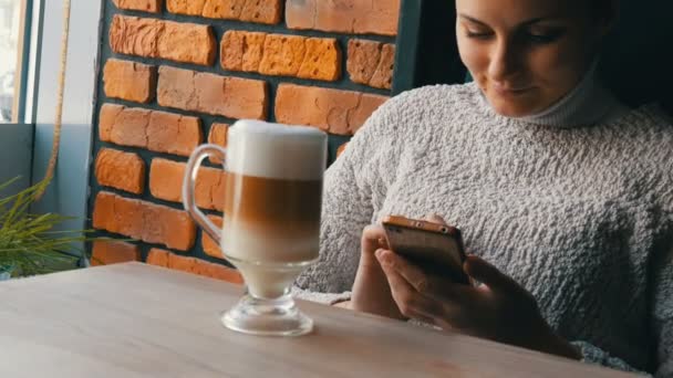 Güzel kız ikindilerde içecekler kahve latte hava köpük ile gülümseyen rahat kafede oturur ve pencereden görünüyor — Stok video