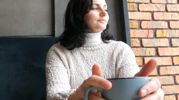 Menina bonita senta-se em um café acolhedor sorrindo pensivamente bebe café e olha pela janela — Vídeo de Stock