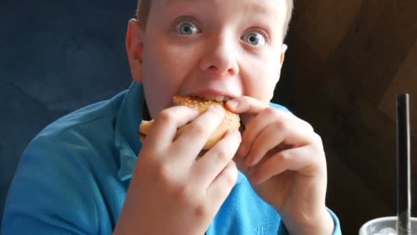 O restaurante fast food, não comida saudável. Azul de olhos bonitos menino adolescente come um grande hambúrguer . — Vídeo de Stock