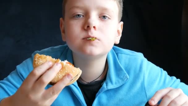 Un guapo adolescente de ojos azules come alternativamente una hamburguesa y alitas de pollo frito en un restaurante de comida rápida y mira una cámara — Vídeo de stock