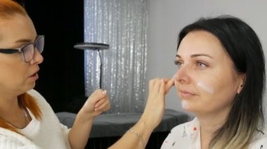 Makyözün yakın plan görüntüsü, makyaj sanatçısı özel bir yüz kremi sürüyor. Mavi gözlü güzel bir kadının yüzünde parmakları var.