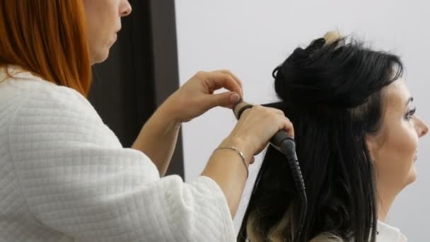 Профессиональный стиль парикмахера делает кудри кудрей с помощью щипцов для завивки волос красивой молодой женщине с длинными волосами окрашенными с использованием комплексной техники в студии красоты — стоковое видео