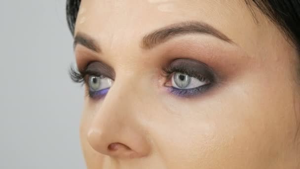 Zblízka pohled na stylista make-up umělce aplikuje nadace krém se speciálním štětcem na tváři mladé krásné ženy s modrýma očima — Stock video