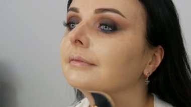 Makyaj sanatçısının yakın plan görüntüsü mavi gözlü güzel bir kadının yüzüne özel bir fırçayla fondöten kremi sürüyor.