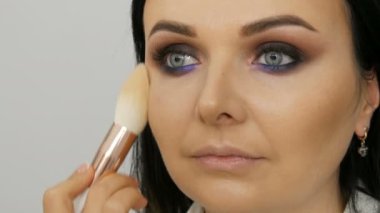 Makyaj sanatçısının yakın plan görüntüsü mavi gözlü güzel bir kadının yüzüne özel bir fırçayla fondöten kremi sürüyor.