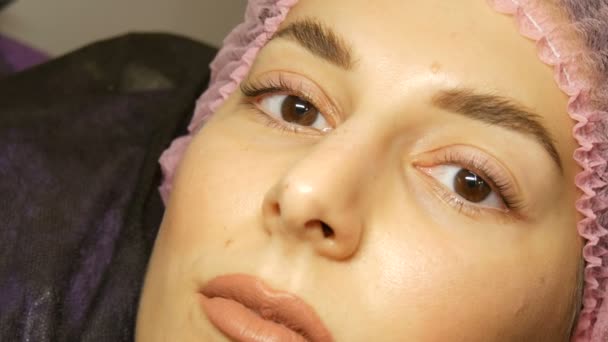 The face of young woman with wide hairy eyebrows and brown eyes in pink hat after the procedure for laminating eyelashes in a beauty salon. Modern eyelash healing — Stock Video