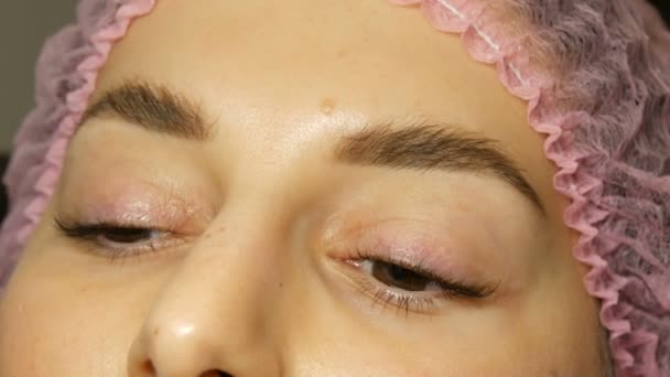 Das Gesicht einer jungen Frau mit breiten behaarten Augenbrauen und braunen Augen in rosa Hut nach der Prozedur zum Laminieren der Wimpern in einem Schönheitssalon. Moderne Wimpernheilkunde — Stockvideo