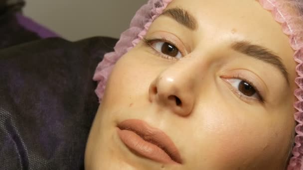 The face of beautiful young woman with wide hairy eyebrows and brown eyes and matte beige lipstick on her lips in a pink hat before the procedure for laminating eyelashes in a beauty salon — Stockvideo