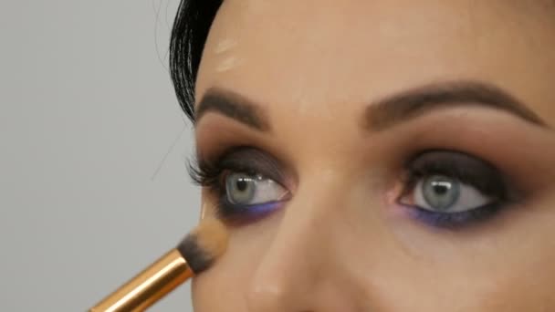 Närbild bild av en stylist makeup artist tillämpar foundation kräm med en speciell borste i ansiktet på en ung vacker kvinna med blå ögon — Stockvideo