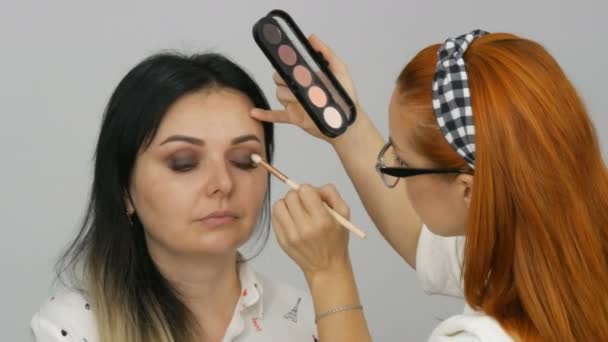 Krásná rusovlasá maskérka dělá make-up pro mladou ženu s béžové oční stíny paleta v salonu krásy — Stock video