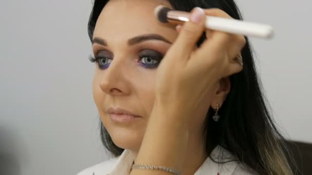 Stylist make-up kunstenaar brengt stichting voor gezicht met een speciale borstel op het gezicht van jonge mooie vrouw met blauwe ogen — Stockvideo