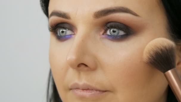 Stylist truccatore applica fondazione per il viso con una spazzola speciale sul viso della giovane bella donna con gli occhi blu — Video Stock