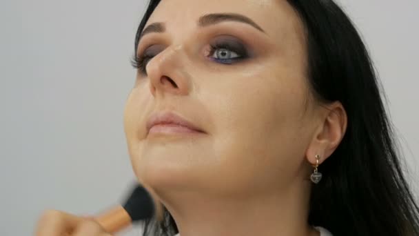 Vista de cerca de un artista de maquillaje estilista aplica crema base con un cepillo especial en la cara de una joven hermosa mujer con ojos azules — Vídeos de Stock