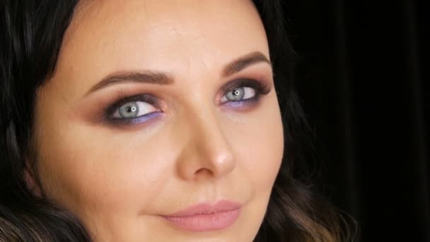 Mujer hermosa joven con ojos azules brillantes y maquillaje de noche ojos ahumados con sombra de ojos azul y púrpura sobre fondo negro — Vídeos de Stock