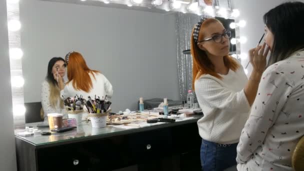 Mooie roodharige make-up kunstenaar doet make-up aan een jonge vrouw met beige oogschaduw palet in een schoonheidssalon — Stockvideo