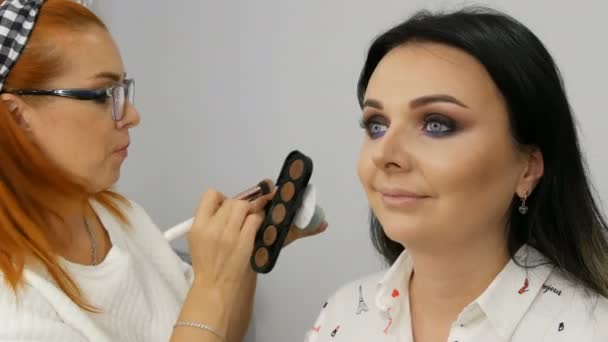 Mooie roodharige make-up kunstenaar doet make-up aan een jonge vrouw met beige oogschaduw palet in een schoonheidssalon — Stockvideo