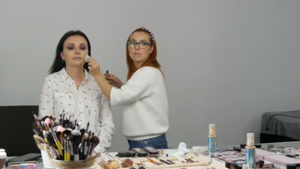 Rothaarige Stylisten Make-up Artist trägt eine spezielle Gesichtscreme mit speziellem Schwamm auf das Gesicht einer jungen schönen Frau mit blauen Augen — Stockvideo