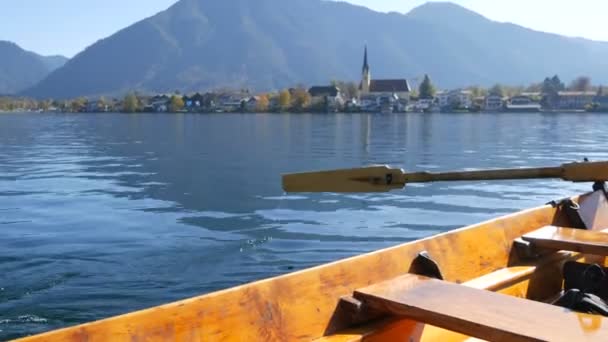 Um barqueiro em barco de madeira com um remo leva as pessoas para o outro lado, contra o fundo de uma bela igreja pitoresca no Lago Tegernsee, Baviera — Vídeo de Stock