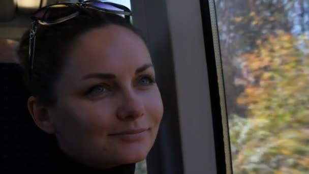 Close-up portret van mooie jonge vrouw met groene ogen en lange zwarte wimpers rijdt in trein kijken dromerig en bedachtzaam uit het raam. Buiten raam is kleurrijke herfst gebladerte. — Stockvideo