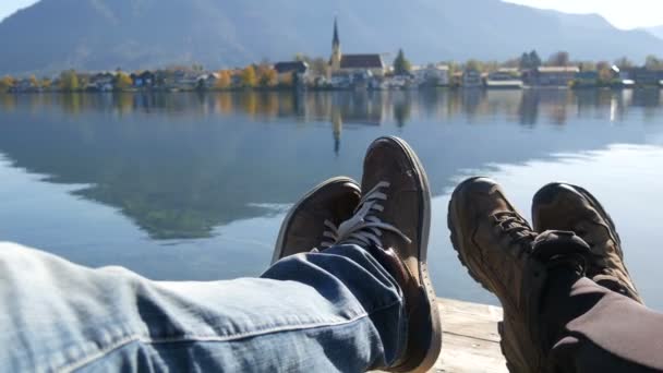 Les voyageurs couple bottes de randonnée sur la montagne près du lac Tegernsee sur le fond de l'église. Chaussures de trekking homme et femme. Paire assis et faire des pieds selfie sur un beau paysage. POV voyager — Video