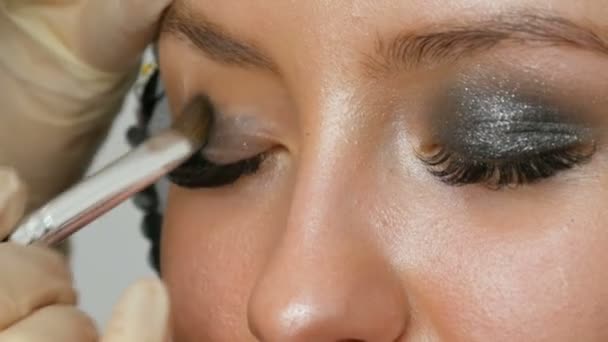 Maquillaje artista hace modelos ojos ahumados con la ayuda de un cepillo especial sombra de ojos gris, ojos y pestañas de niña vista de cerca. Profesional de alta moda . — Vídeos de Stock