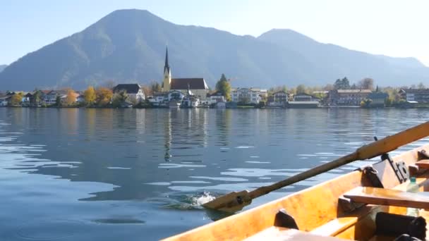 Una imagen tranquila un barco de madera con un remo flota en el hermoso lago de montaña Tegernsee contra el telón de fondo de las montañas alpinas y el pintoresco contenedor de la iglesia. Ferryman transbordadores personas — Vídeo de stock