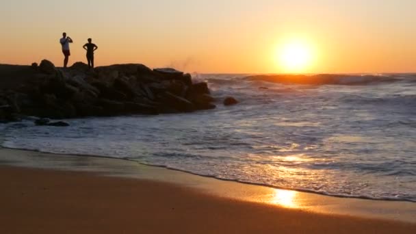 Salida del sol o puesta de sol muy hermosa en la orilla del mar. Playa de arena y olas que golpean en la playa. Grandes olas de tormenta con espuma. Siluetas de un par de personas caminando sobre un acantilado rocoso y mirando al mar — Vídeo de stock