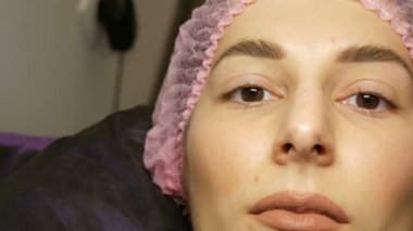 Geniş kıllı kaşları, kahverengi gözleri ve pembe şapkasıyla dudaklarında mat bej ruj izi olan güzel bir kadının yüzü güzellik salonunda kirpikleri kaplama işleminden önce.