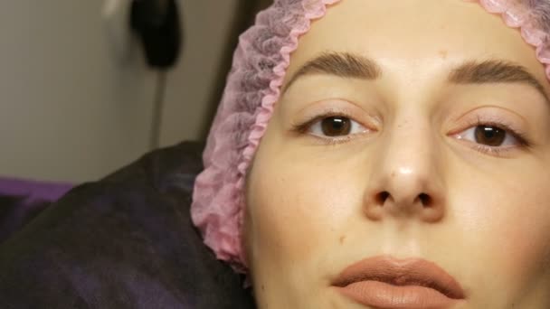 The face of beautiful young woman with wide hairy eyebrows and brown eyes and matte beige lipstick on her lips in a pink hat before the procedure for laminating eyelashes in a beauty salon — Stockvideo