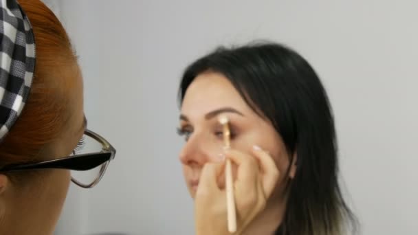 Ansikte på vackra rödhåriga makeup artist i glasögon gör makeup till en ung kvinna i skönhetssalong — Stockvideo