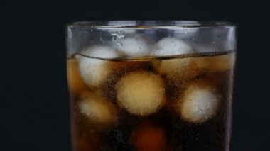 Soğuk kola kabarcıkları şeffaf uzun bir bardakta, siyah bir arkaplanda dönen buzla yakın plan görüntüsü var..
