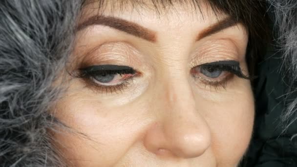 Ενηλίκων μεσήλικη γυναίκα με όμορφα μεγάλα μπλε μάτια, φωτεινό μακιγιάζ, βαμμένα βέλη και ψεύτικες βλεφαρίδες σε μια ψεύτικη γούνα κουκούλα σε λευκό φόντο — Αρχείο Βίντεο