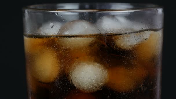 Bubbles of cold cola in a transparent long glass with ice on a black background close up view. — Stock Video