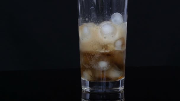 Cola se vierte en una gran taza de vidrio largo con trozos de hielo en el interior sobre un fondo negro . — Vídeo de stock