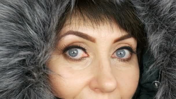 Mujer adulta de mediana edad con hermosos ojos azules grandes, maquillaje brillante, flechas pintadas y pestañas falsas en una capucha de piel sintética sobre fondo blanco — Vídeos de Stock