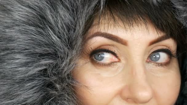 Mujer adulta de mediana edad con hermosos ojos azules grandes, maquillaje brillante, flechas pintadas y pestañas falsas en una capucha de piel sintética sobre fondo blanco — Vídeo de stock