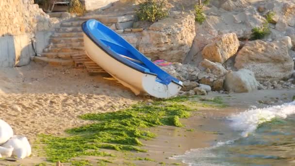 Barco de pesca azul y blanco vacío en la orilla del mar en el que se lanzan algas verdes después de una tormenta. Olas con espuma golpean contra la orilla — Vídeos de Stock