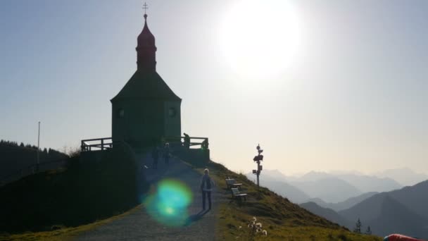 Tegernsee, Németország - 2019. október 23.: Régi templom a bajor Alpok festői lejtőjén, amely közelében a turisták sétálnak, hegyi siklóernyőzés — Stock videók
