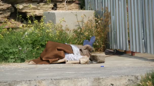 Homem sem-teto mente e dorme na rua sob cerca coberta com material do sol — Vídeo de Stock