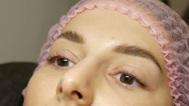 Geniş kıllı kaşları, kahverengi gözleri ve pembe şapkasıyla dudaklarında mat bej ruj izi olan güzel bir kadının yüzü güzellik salonunda kirpikleri kaplama işleminden önce.