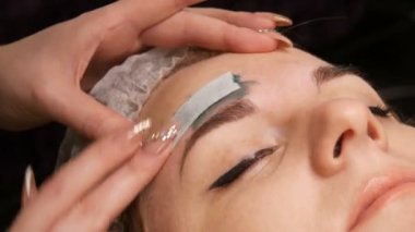 Kaşların şeklinin balmumu düzeltmesi. Güzellik uzmanı kaşlarına sıcak yeşil balmumu sürüyor ve saçlarını özel bir şeritle temizliyor..