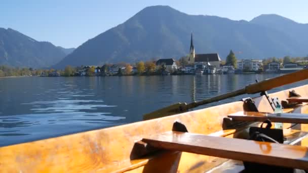 Una imagen tranquila un barco de madera con un remo flota en el hermoso lago de montaña Tegernsee contra el telón de fondo de las montañas alpinas y el pintoresco contenedor de la iglesia. Ferryman transbordadores personas — Vídeos de Stock